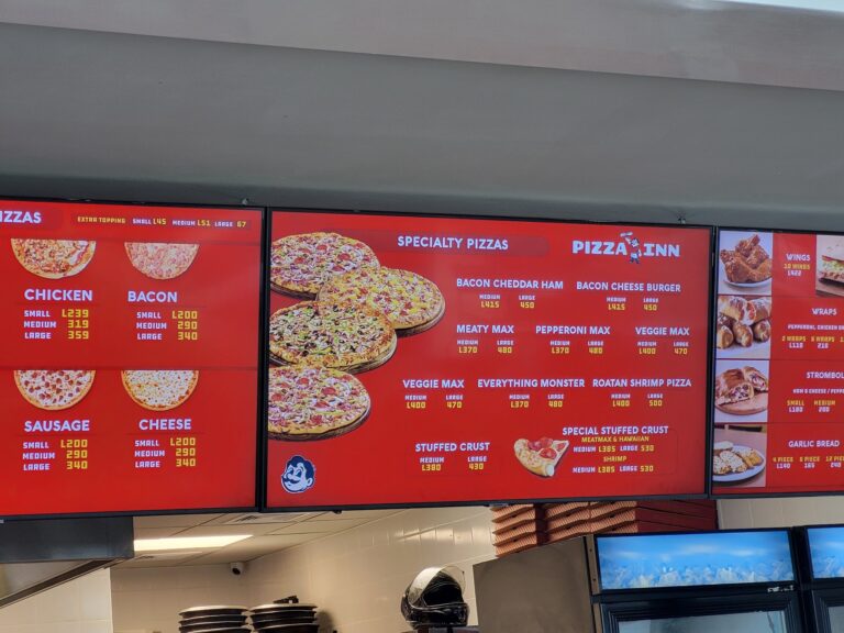 A digital menu board at a pizza restaurant in Roatan, Honduras, displaying various specialty pizzas, toppings, and prices.