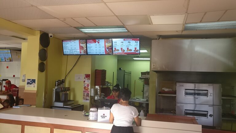 Inside a fast-food restaurant on Roatan, Honduras, staff members prepare orders behind the counter while digital menu screens display pizza and combo options in a brightly lit, casual setting.