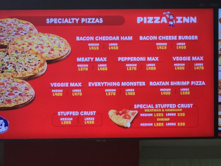 A bright red digital menu board for Pizza Inn in Roatan, Honduras, displays various specialty pizzas such as bacon cheddar ham, bacon cheese burger, veggie max, and shrimp, with prices for medium and large sizes.