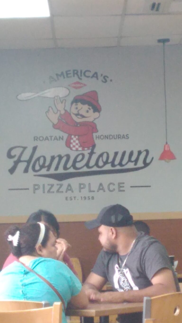 People are sitting and talking inside Hometown Pizza Place in Roatan, Honduras, with a vintage-style mural of a cheerful cartoon boy holding a pizza and the restaurant's name on the wall.