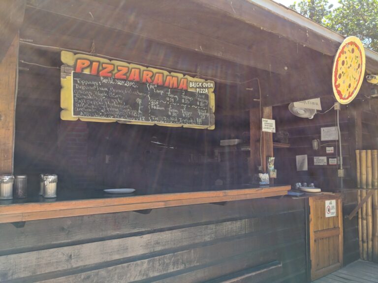 A wooden wall with a large colorful pizza sign and a chalkboard menu advertising pizza options at a restaurant in Roatan, Honduras.