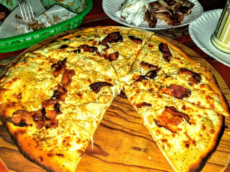 A freshly baked pepperoni and cheese pizza sits on a wooden platter surrounded by plates of grilled chicken wings, set on a table at a restaurant in Roatan, Honduras.
