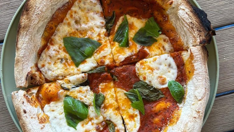 A Margherita pizza with fresh basil leaves on a green plate, featuring a golden crust and melted cheese, served on a wooden table in Roatan, Honduras.