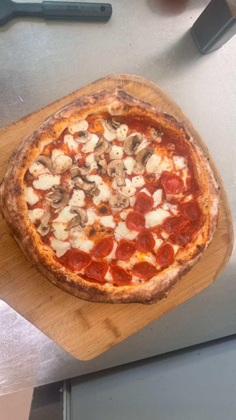 A pepperoni and mushroom pizza with melted cheese on a wooden serving board, placed on a stainless steel table in Roatan, Honduras.