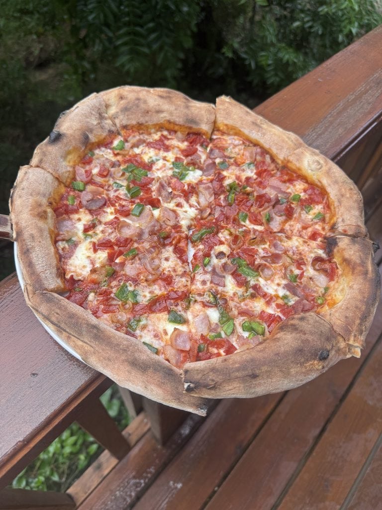 A freshly baked pizza with toppings of tomatoes, green onions, and ham sits in a wooden pizza peel on a wooden table outdoors in Roatan, Honduras.