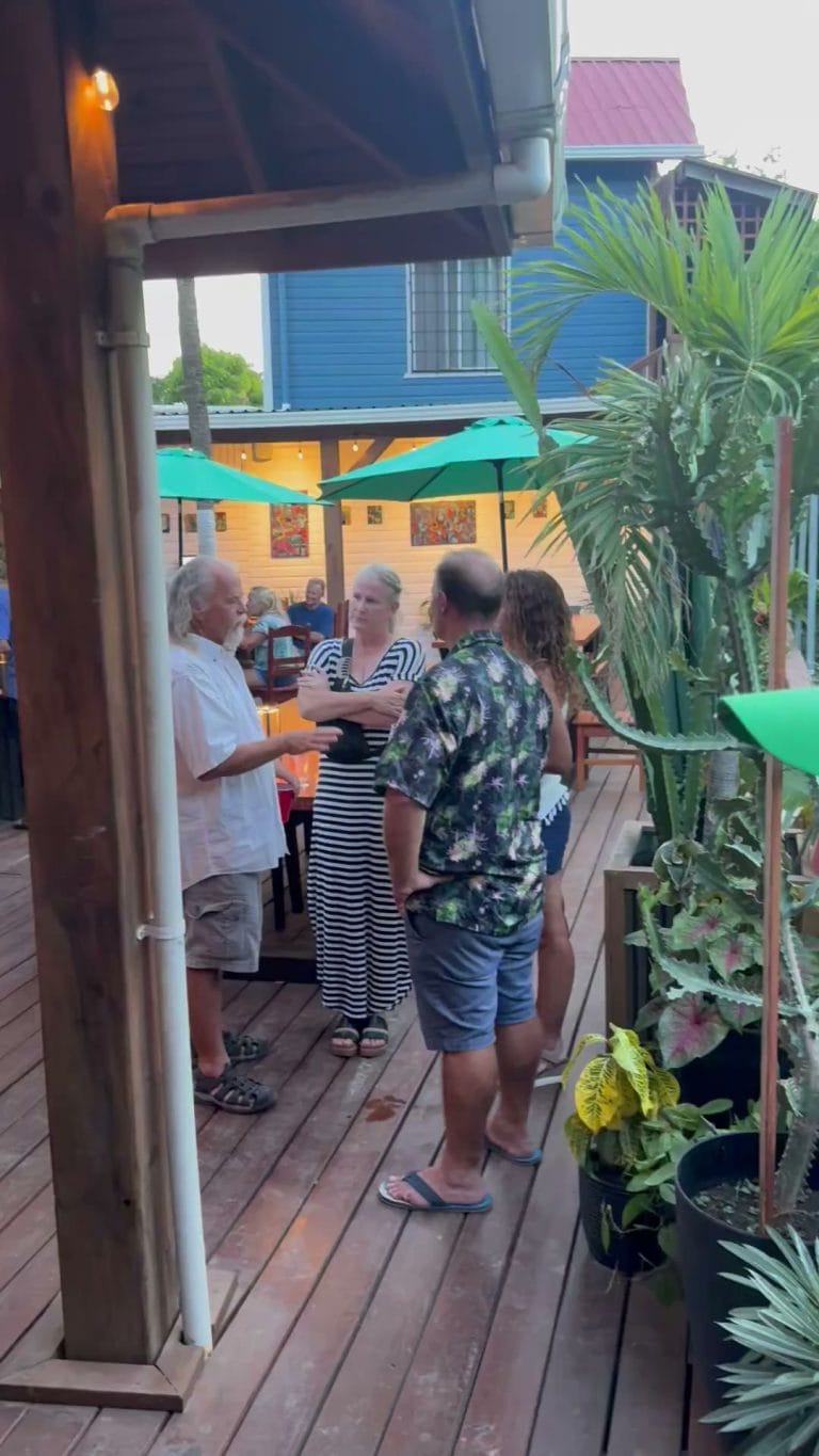 People are gathered and talking on a wooden deck patio surrounded by tropical plants, with colorful umbrellas and a blue house in the background in Roatan, Honduras.