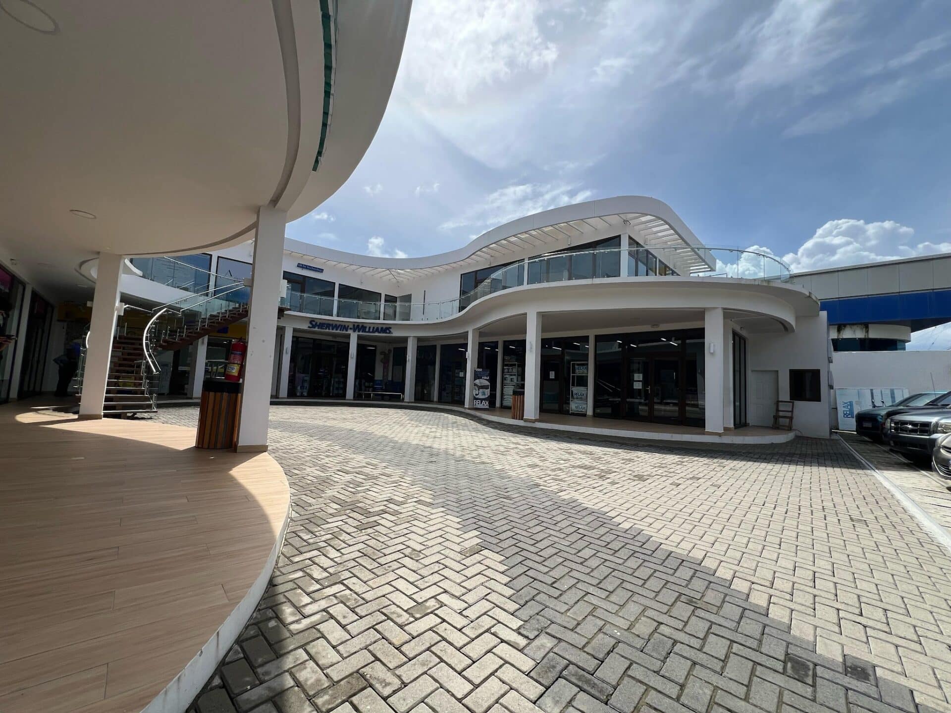 A modern, white commercial building with curved architectural design and glass balcony railings located in Roatan, Honduras, featuring a paved outdoor area with brick and wooden flooring under a partly cloudy sky.