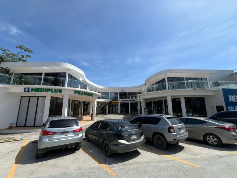 Modern two-story shopping center with glass balconies, featuring a pharmacy named MEDIPLUS and other retail stores, located in Roatan, Honduras, with parked cars in the foreground and a clear blue sky overhead.