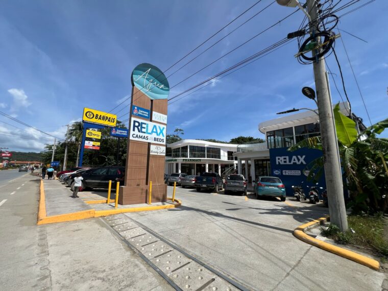 A commercial area in Roatan, Honduras, featuring a parking lot with parked cars, a prominent sign for "Relax" offering beds and a salon, and modern white buildings housing a pharmacy and retail stores under a clear blue sky.