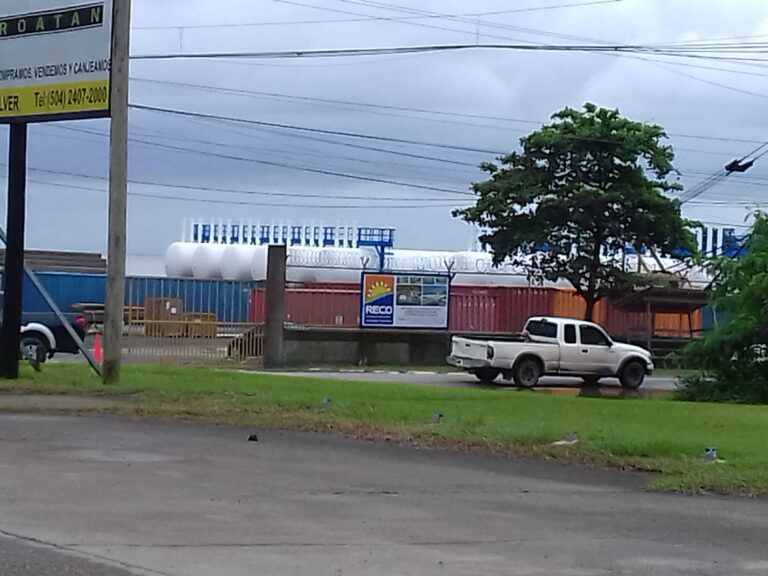 A street view in Roatan, Honduras shows a large sign with the words "Roatan" and a gas station or industrial facility with tanks and metal structures in the background, alongside utility poles and vehicles parked along the road.