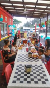 A family gathers around a long checkered table with red chairs on a covered patio decorated with blue and white balloons, enjoying a meal in Roatan, Honduras.
