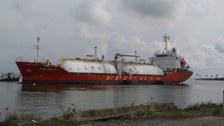 A large red and white LPG gas tanker named "EPIC BALTA" is docked in Roatan, Honduras, with palm trees and a cloudy sky visible in the background.