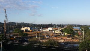 A panoramic view of a construction site and small buildings near the water in Roatan, Honduras, with hills and a partly cloudy sky in the background.