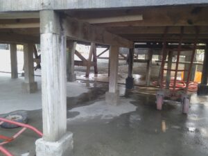 A wooden building under construction on Roatan, Honduras, with visible support beams, concrete footings, and construction materials like a ladder, hoses, and tires on the ground.
