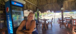 A smiling woman sits at a wooden table inside a bar with a thatched roof in Roatan, Honduras, featuring a Pepsi refrigerator, wooden chairs, and a view of parked cars outside.