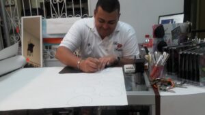 A man working at a cluttered desk in an office in Roatan, Honduras, is writing or drawing on a sheet of paper surrounded by office supplies, electronics, and personal items.