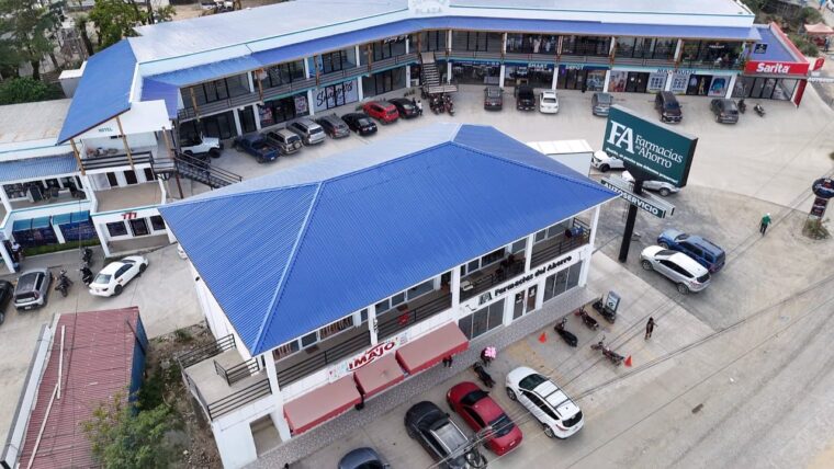 An aerial view of a commercial building with a bright blue metal roof on Roatan, Honduras, featuring multiple shops, a parking lot with cars and motorcycles, and a large sign for Farmacias Ahorro in a busy area of the island.