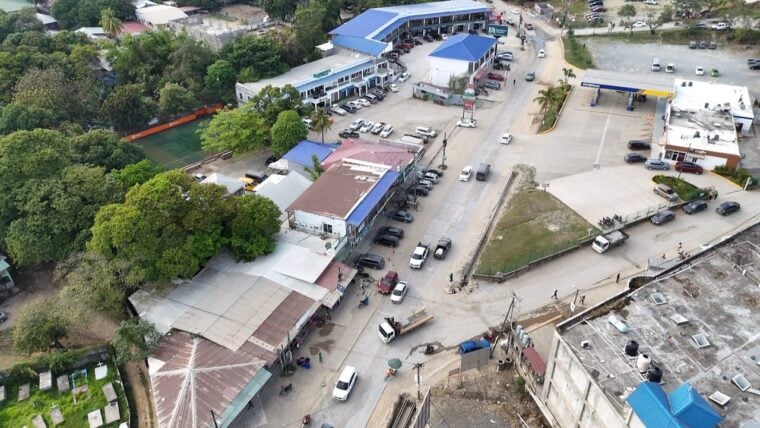 An aerial view of a commercial area in Roatan, Honduras, featuring a parking lot, businesses with colorful roofs, surrounding greenery, and a tennis court amidst the town infrastructure.