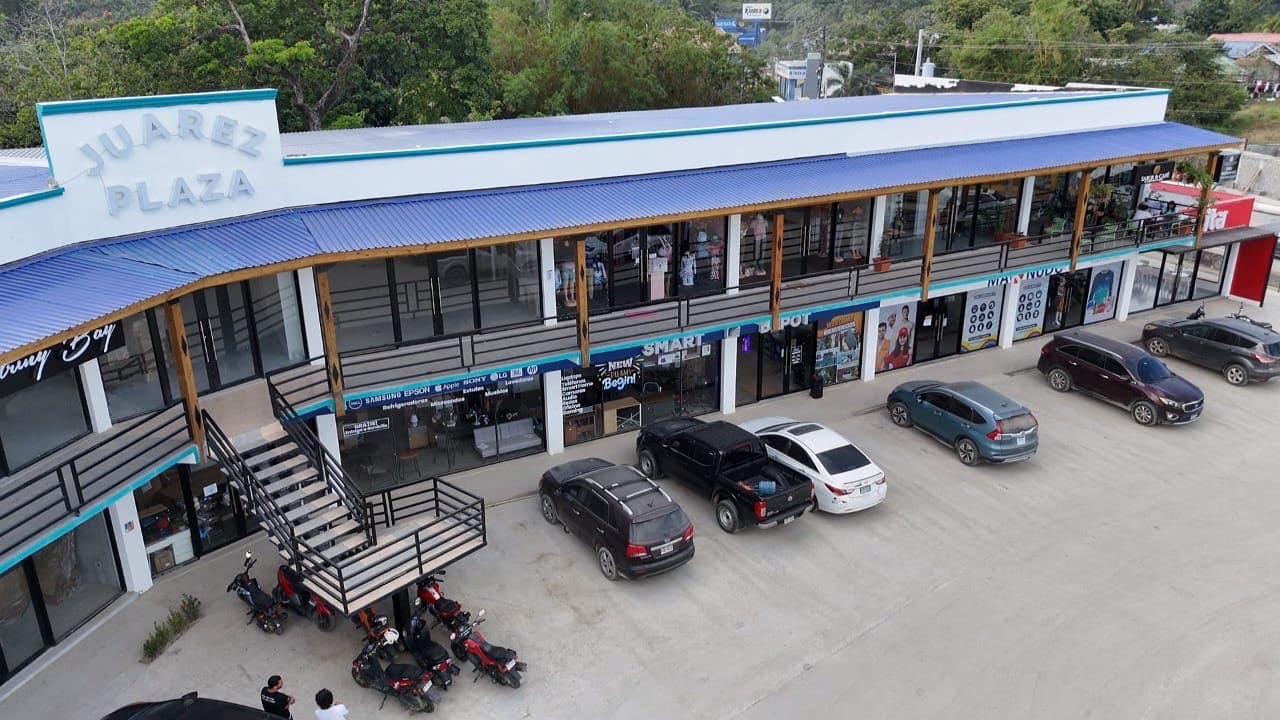 Juares Plaza, a two-story shopping and commercial center in Roatan, Honduras, features storefronts with large glass windows, a blue metal roof, an outdoor staircase, and parking spaces with several cars and motorcycles in front.