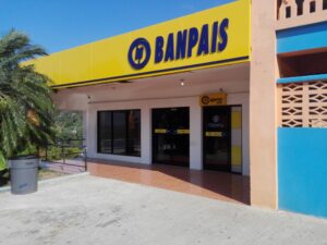 A storefront in Roatan, Honduras, features a prominent yellow sign with blue lettering for Banpais, a financial services business, with a palm tree on the left and a blue sky overhead.