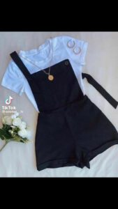 A casual outfit featuring a white t-shirt layered under black overalls with one strap hanging, accessorized with a gold necklace and hoop earrings, displayed on a white surface next to a small bouquet of white flowers, in a business listing on RoatanYP.com in Honduras.