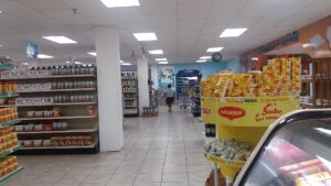 The interior of a supermarket in Roatan, Honduras, displaying shelves stocked with various food products, including a prominent round display of Maggi noodles and customers shopping in a well-lit space with tropical-themed decorations on the wall.