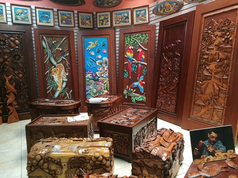 A display of intricately carved wooden artwork and jewelry featuring tropical motifs, with colorful paintings of marine life and parrots on the wall in a Roatan, Honduras souvenir shop.