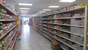 A grocery store aisle in Roatan, Honduras, displays shelves stocked with various food products, including canned goods and snacks, with a customer shopping near the window at the far end.