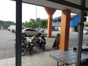 A view from inside a storefront showing motorcycles parked outside, colorful buildings with blue and orange facades, lush green trees, and a partly cloudy sky in Roatan, Honduras.