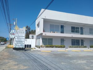 A modern white two-story business building in Roatan, Honduras, with a glass balcony on the upper floor, surrounded by a small landscaped area with shrubs and plants, and a signage board displaying various company names including Insura, Apolo Group, and Mouv near the street.