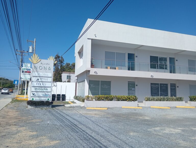 A modern white two-story business building in Roatan, Honduras, with a glass balcony on the upper floor, surrounded by a small landscaped area with shrubs and plants, and a signage board displaying various company names including Insura, Apolo Group, and Mouv near the street.