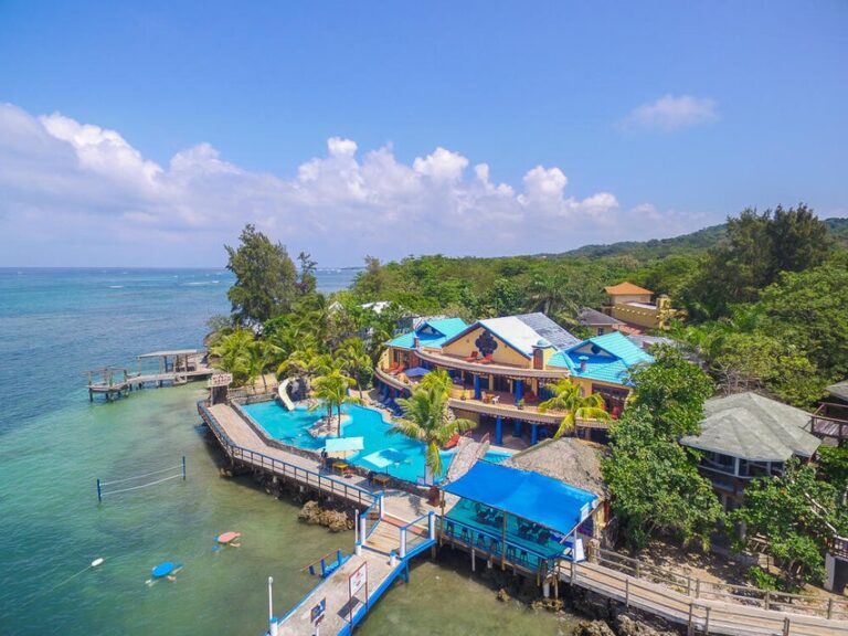 Colorful beachfront resort with blue-roofed buildings, palm trees, and a swimming pool on Roatan, Honduras overlooking the Caribbean Sea.