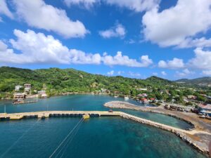 A scenic view of a harbor in Roatan, Honduras, featuring a marina with boats, a curved breakwater, lush green hills with residential buildings, and a partly cloudy blue sky.