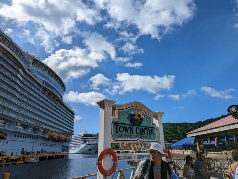 People walk along the waterfront at Roatan in Honduras, with large cruise ships docked nearby and a sign for the Town Center at Port of Roatan in the foreground.