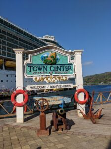 A decorative sign reading "Town Center at Port of Roatan" is displayed at the harbor in Roatan, Honduras, surrounded by nautical-themed props such as life preservers, a ship's wheel, and an anchor, with a cruise ship docked nearby and a hillside visible in the background.