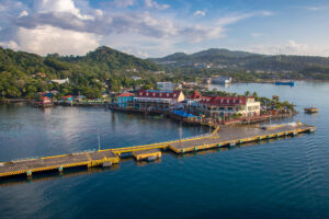 A view of Roatan's waterfront in Honduras showing colorful buildings, a dock with boats, lush green hills, and a partly cloudy sky.