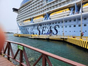 A large cruise ship docked in Roatan, Honduras, featuring multiple yellow lifeboats and a waterfront walkway with a red railing and greenery in the background.