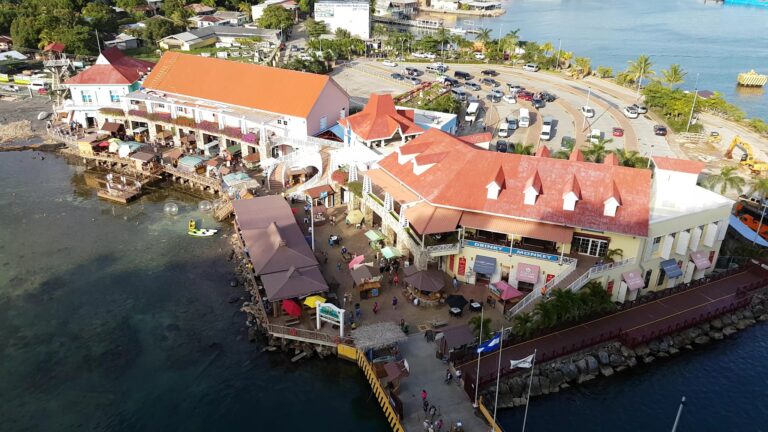 Aerial view of a bustling waterfront shopping and dining area in Roatan, Honduras, featuring colorful buildings, outdoor patios, and a parking lot along the coast with lush greenery and calm blue waters.