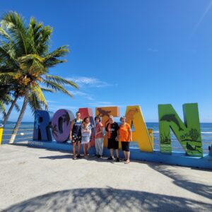 A group of five people stands smiling in front of colorful large letters spelling "ROATAN" along the beachfront in Roatan, Honduras, with a palm tree, clear blue sky, and ocean in the background.