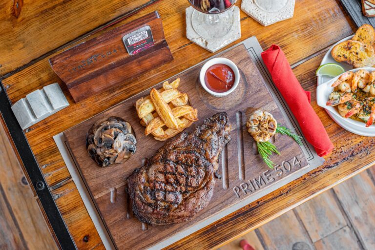 A wooden table displays aServing platter of grilled steak, French fries with ketchup, sautéed mushrooms, a bowl of shrimp in sauce, a slice of toast, and slices of lime, with a glass of red wine and a red cloth napkin, at a restaurant in Roatan, Honduras.