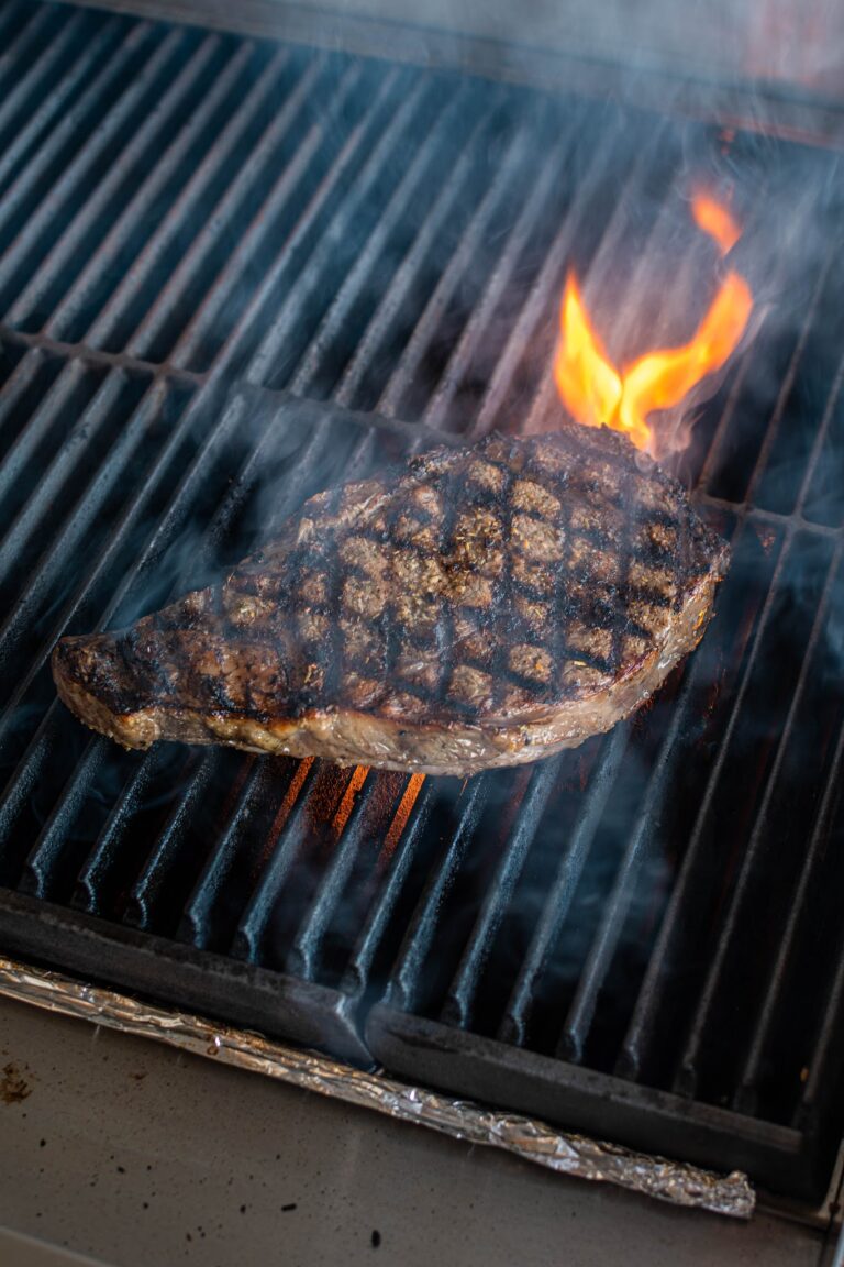 A seasoned steak is grilling over an open flame on a barbecue in Roatan, Honduras, with visible grill grates and smoke rising around the meat.