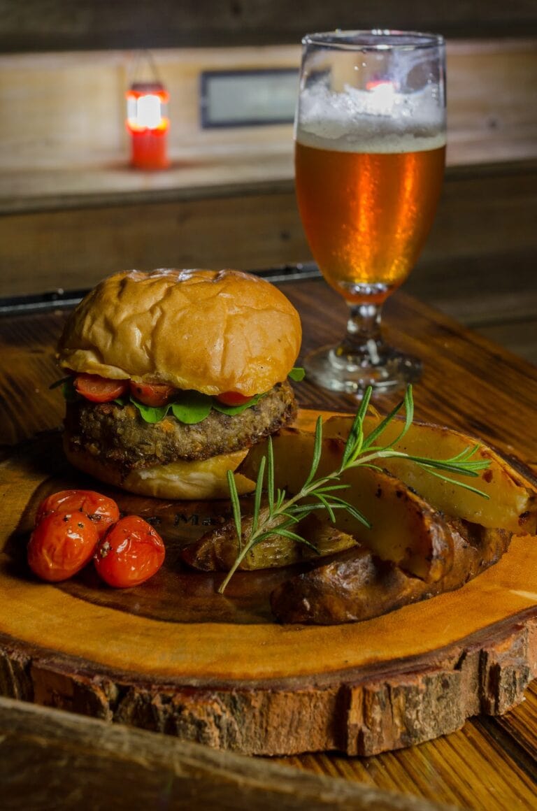 A burger with fresh vegetables and roasted potatoes is served on a wooden platter in a restaurant setting in Roatan, Honduras, with a glass of beer visible in the background.