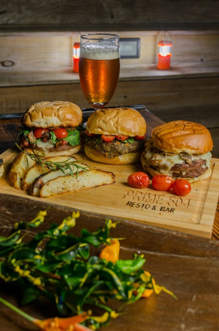 A wooden serving board with three gourmet burgers topped with fresh vegetables and melted cheese, accompanied by roasted potato slices, with a glass of amber-colored beer behind, set against a rustic wooden background with red lamps in a Roatan, Honduras restaurant or bar.