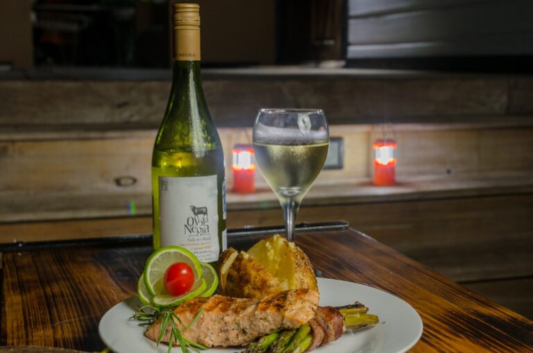 A plate of grilled salmon served with lemon slices, cherry tomatoes, and asparagus is shown in front of a bottle of white wine and a glass of wine, with a rustic wooden table and soft ambient lighting in a Roatan restaurant in Honduras.