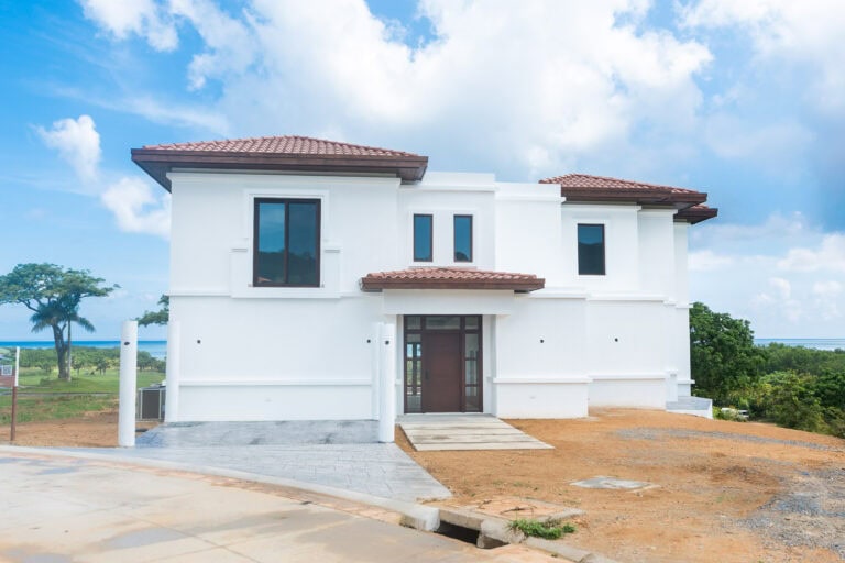 New white two-story house with a red-tiled roof situated on a hilltop in Roatan, Honduras, overlooking lush greenery and distant ocean views under a partly cloudy sky.
