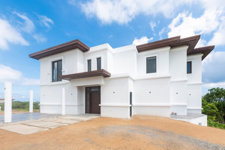 Modern white two-story house with dark brown roof accents located on Roatan in Honduras, with a clear blue sky and lush greenery in the background.