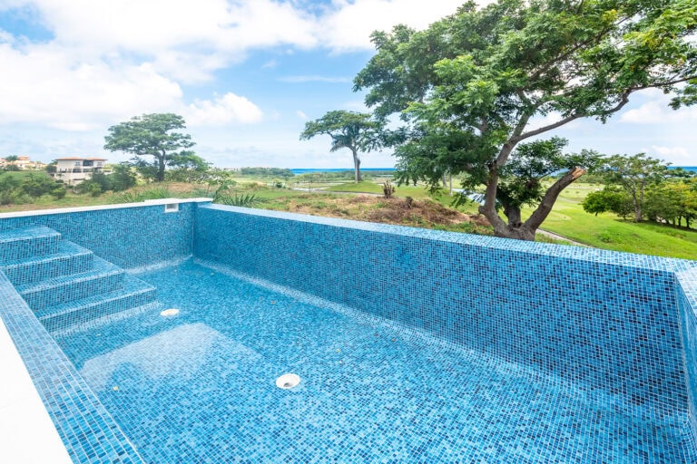 A private blue-tiled swimming pool overlooking grassy fields, trees, and the turquoise waters of Roatan in Honduras under a partly cloudy sky.