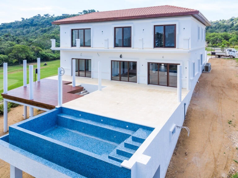 A modern, white three-story house on Roatan in Honduras with large glass doors and windows, an outdoor terrace with wooden deck, and a blue tiled swimming pool with stairs, set against lush green hills.