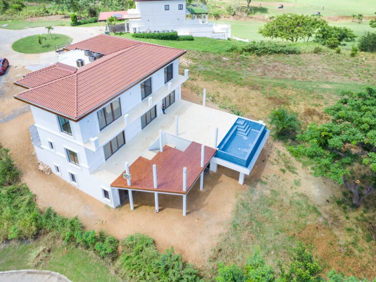A white three-story building with a red-tiled roof facing the ocean in Roatan, Honduras, featuring large glass windows and an outdoor terrace with a pool and wooden deck.