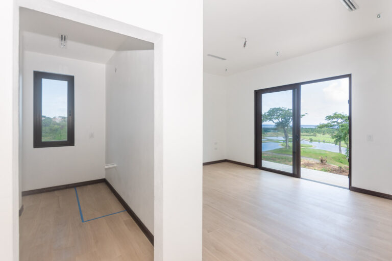 Interior view of a modern, unfurnished living room in a house on Roatan, Bay Islands, Honduras, with large glass sliding doors overlooking a lush green golf course and water feature.